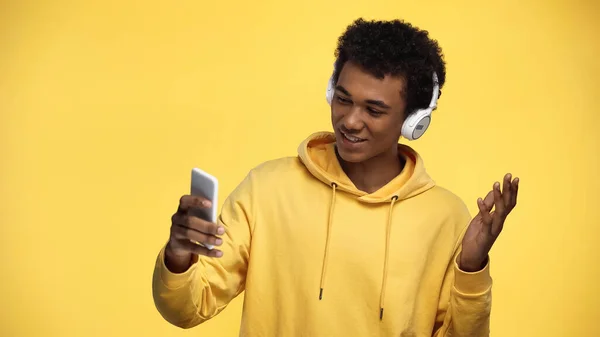 Happy african american teenage boy in wireless headphones using smartphone isolated on yellow — Stock Photo