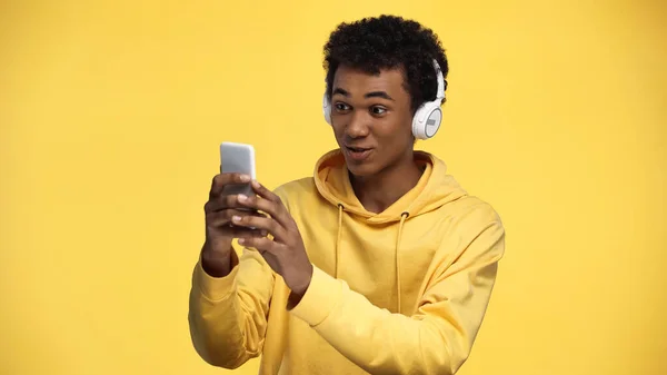 Cheerful african american teenage boy in wireless headphones using smartphone isolated on yellow — Stock Photo