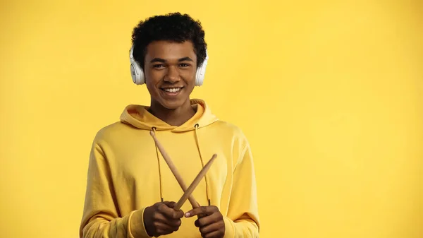 Cheerful african american teenager in wireless headphones holding drumsticks isolated on yellow — Stock Photo