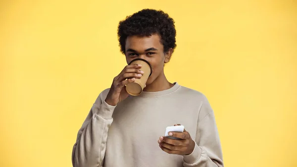 Afroamericano adolescente en sudadera bebiendo café para ir y sosteniendo teléfono inteligente aislado en amarillo - foto de stock