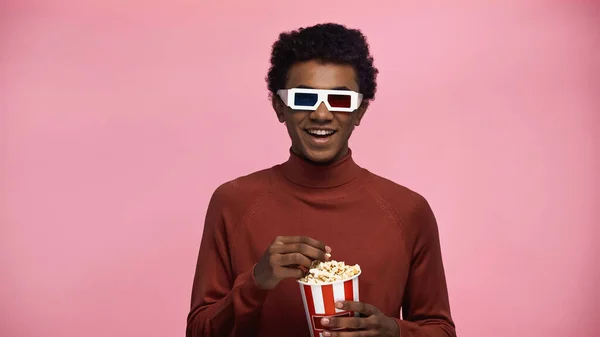 Happy african american teenager in 3d glasses holding popcorn bucket isolated on pink — Stock Photo
