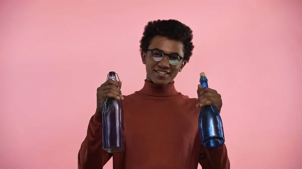 African american teenager in turtleneck sweater and eyeglasses looking at camera while holding bottles isolated on pink — Stock Photo