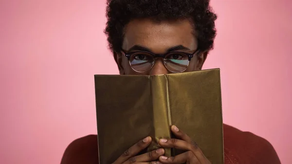 African american teenager obscuring face with book isolated on pink — Stock Photo