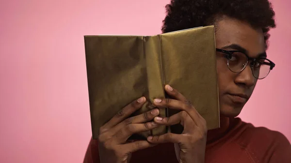 African american teenager holding book in golden cover isolated on pink — Stock Photo