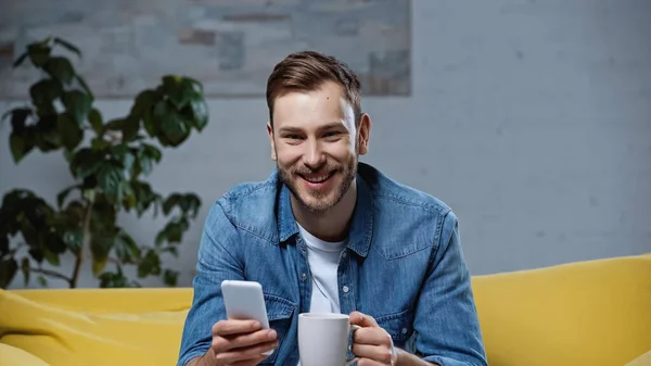 Homem alegre segurando smartphone e xícara de café na sala de estar — Fotografia de Stock