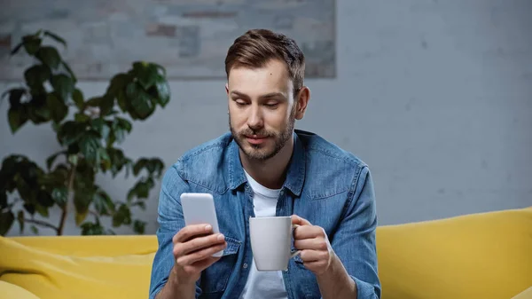 Homem barbudo usando smartphone e segurando xícara de café na sala de estar — Fotografia de Stock