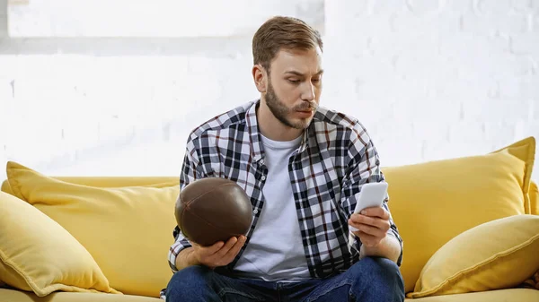 Американский футбольный фанат держит регби мяч и смотрит матч на смартфоне — стоковое фото