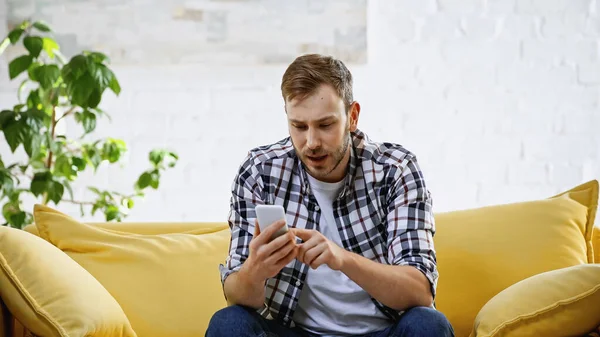 Young man in checkered shirt sitting on sofa and looking at smartphone — Stock Photo