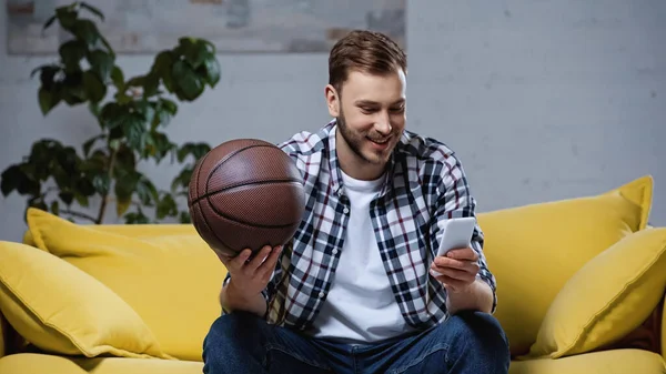 Счастливый баскетбольный болельщик держит мяч и смотрит матч на смартфоне — стоковое фото