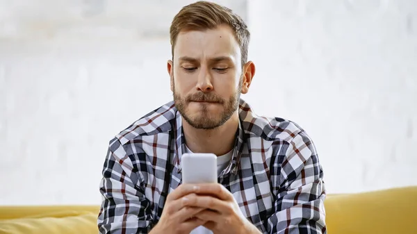 Homem barbudo na camisa quadriculada mordendo lábios enquanto mensagens no smartphone — Fotografia de Stock