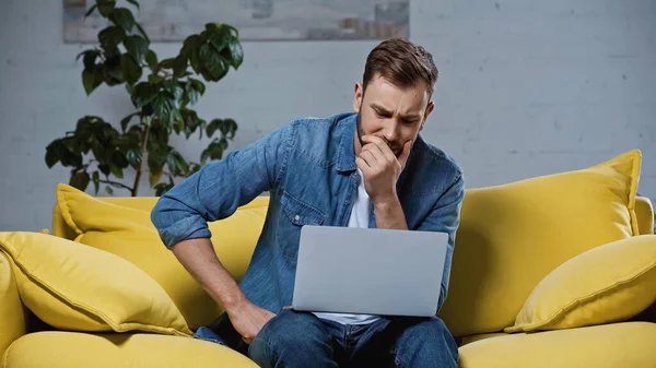 Sleepy freelancer sitting on sofa, yawning and using laptop — Stock Photo