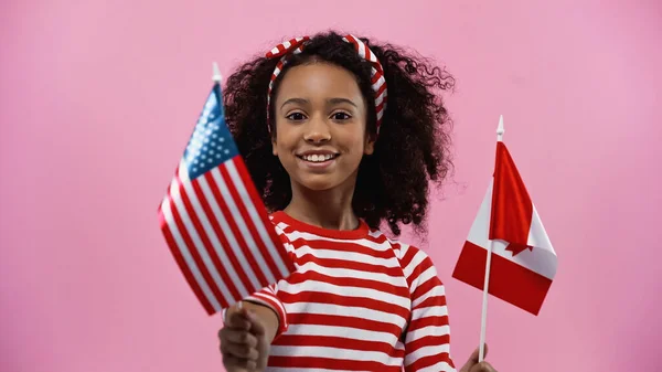 Feliz afroamericana chica sosteniendo banderas de america y canada aislado en rosa - foto de stock