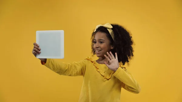 Feliz afroamericano chica saludando la mano mientras que tiene videollamada en tableta digital aislado en amarillo - foto de stock