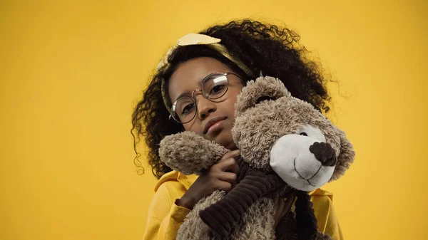 Кучерява афроамериканська дівчина в окулярах тримає плюшевого ведмедя ізольовано на жовтому — стокове фото