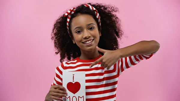 Happy african american girl pointing with finger at greeting card with i love mom lettering isolated on pink — Stock Photo