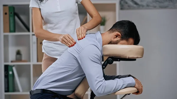 Masajista masajeando espalda de hombre de negocios en silla de masaje en la oficina - foto de stock