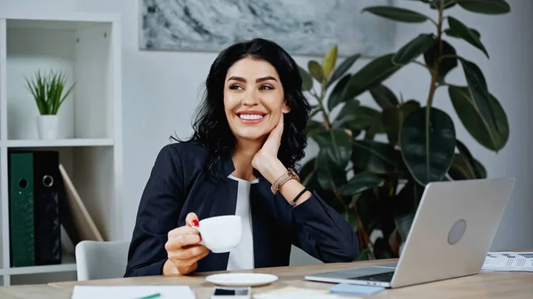 Mujer de negocios sonriente sosteniendo la taza cerca de dispositivos en primer plano borroso - foto de stock