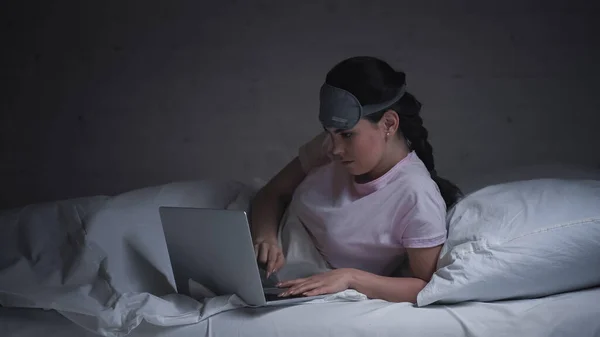 Young freelancer in eye mask using laptop in bed at night — Stock Photo