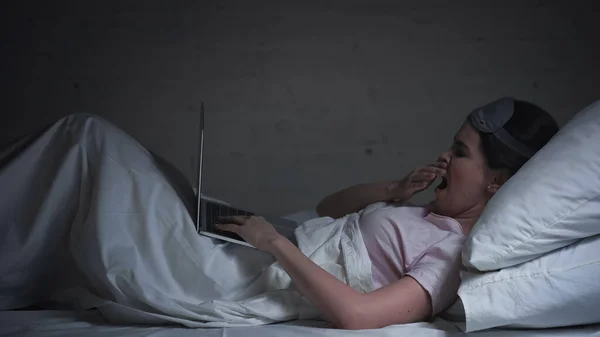 Tired freelancer in eye mask using laptop and yawning in bed at night — Stock Photo