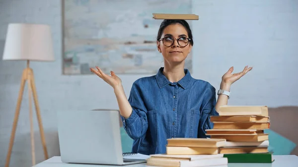 Giovane donna in occhiali con libro sulla testa vicino al computer portatile sulla scrivania — Foto stock