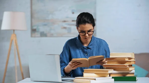 Giovane donna in occhiali lettura libro vicino computer portatile sulla scrivania — Foto stock