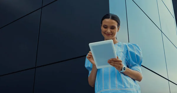 Vue à angle bas de la femme heureuse en chemisier en utilisant une tablette numérique tout en se tenant près du bâtiment — Photo de stock