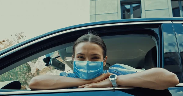 Young woman in medical mask sitting on driver seat and looking at camera from open window — Stock Photo