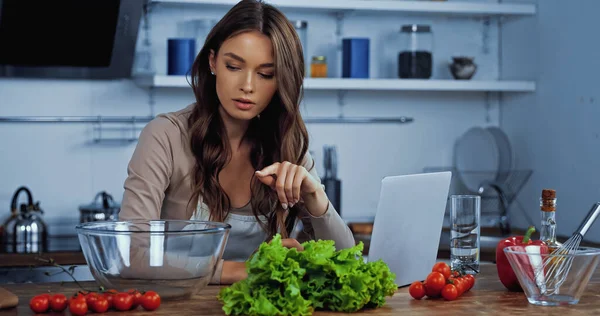 Молода жінка вказує пальцем на інгредієнти біля ноутбука на столі — Stock Photo