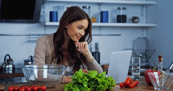 Happy woman searching recipe in laptop near ingredients on table — Stock Photo