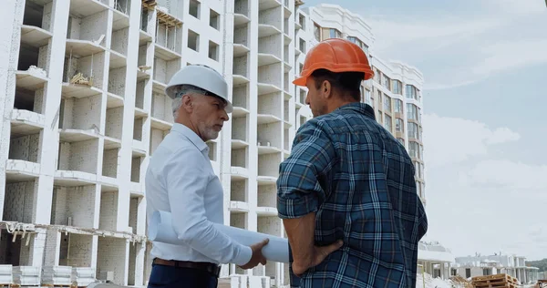 Engineer with blueprints talking with builder standing with hand on hip on construction site — Stock Photo