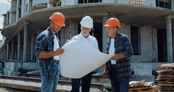 Engineer and builders in hard hats discussing blueprint on construction site — Stock Photo