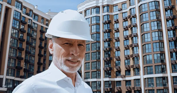 Middle aged engineer looking at camera while smiling near building on background — Stock Photo