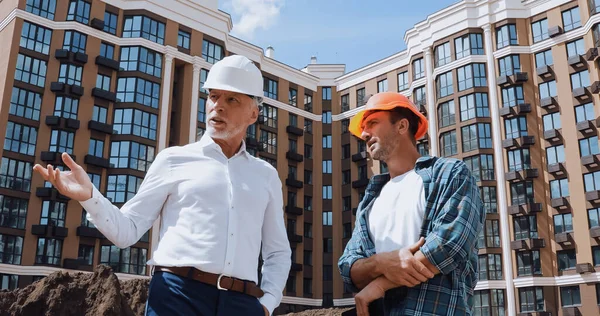 Vista de ángulo bajo de ingeniero gesto cerca constructor mientras habla cerca de nuevo edificio — Stock Photo