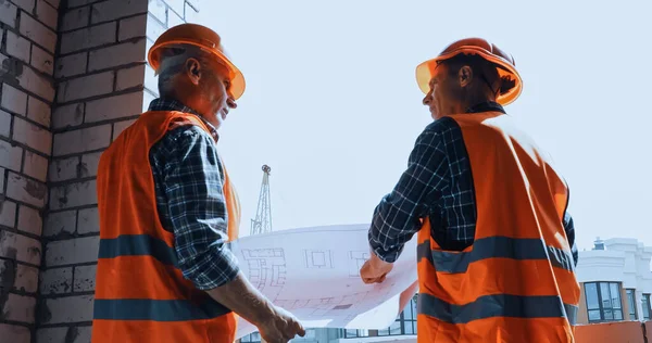 Low angle view of builders in safety vests with blueprint talking on construction site — Stock Photo
