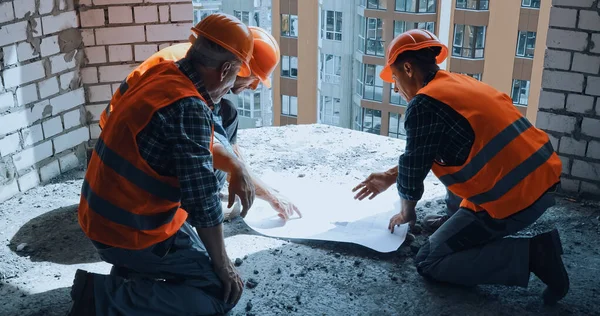Três construtores em chapéus de mão discutindo o projeto enquanto sentados no canteiro de obras — Fotografia de Stock