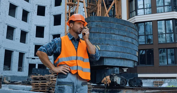 Builder standing with hand on hip and using walkie talkie on construction site — Stock Photo
