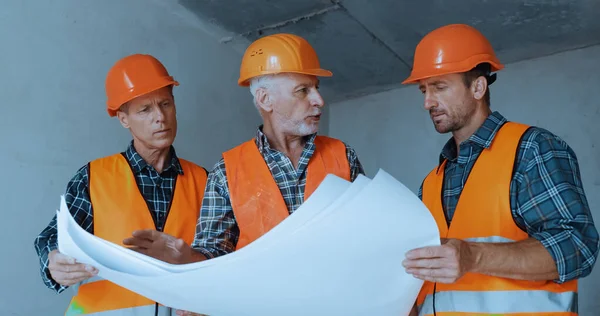 Construtores em capacetes de segurança falando enquanto seguram plantas no canteiro de obras — Fotografia de Stock
