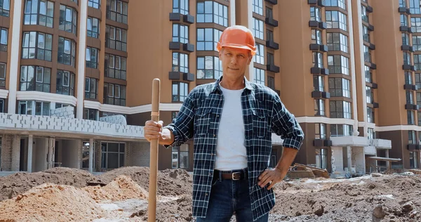 Constructor sonriente sosteniendo la pala y de pie con la mano en la cadera en el sitio de construcción - foto de stock