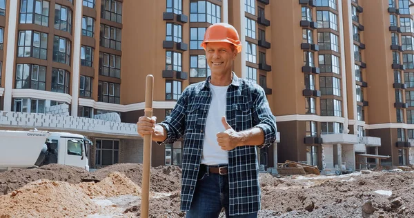 Lächelnder Bauarbeiter hält Schaufel und zeigt Daumen nach oben auf Baustelle — Stockfoto