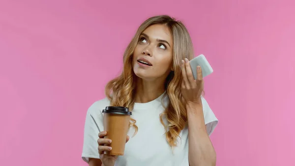 Curious woman using smartphone while listening voice message and holding coffee to go isolated on pink — Stock Photo