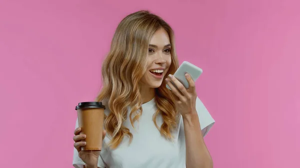 Blonde woman using smartphone while recording voice message and holding coffee to go isolated on pink — Stock Photo