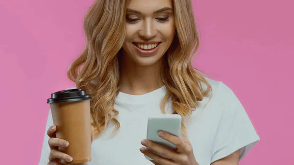 Веселая женщина с помощью смартфона и проведение кофе, чтобы пойти изолированы на розовый — стоковое фото