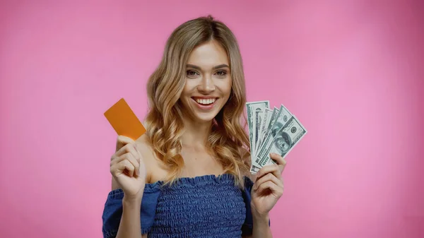 Alegre mujer rubia sosteniendo tarjeta de crédito y dólares aislados en rosa - foto de stock