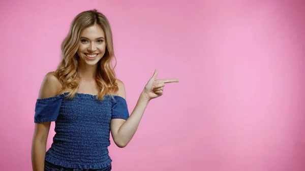 Mujer rubia alegre señalando con el dedo y mirando a la cámara aislada en rosa - foto de stock