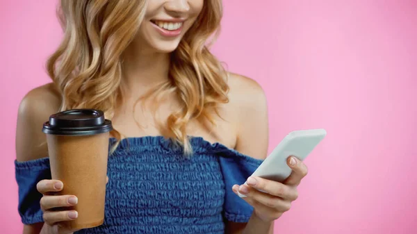Vista recortada de la mujer alegre usando smartphone y sosteniendo café para ir aislado en rosa - foto de stock