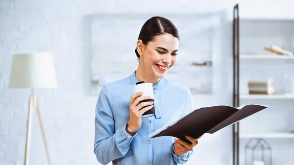 Cheerful businesswoman holding coffee to go and folder with documents in office — Stock Photo
