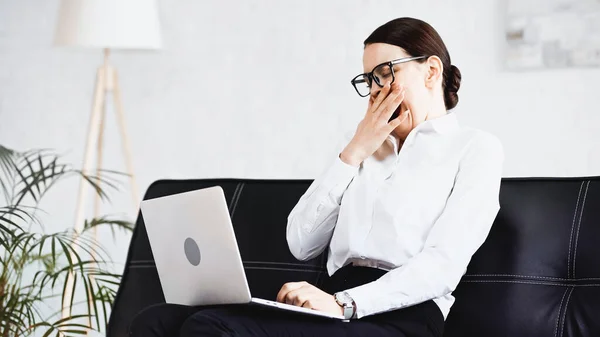 Sleepy businesswoman yawning while sitting on sofa with laptop — Stock Photo