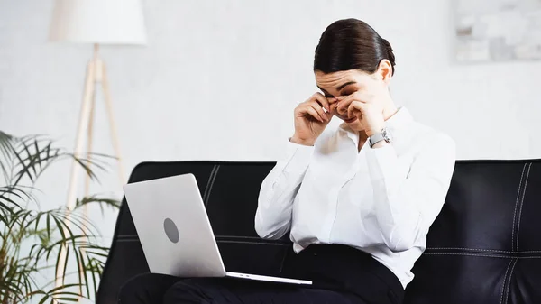 Mujer de negocios agotado tocar los ojos mientras se sienta en el sofá con el ordenador portátil - foto de stock