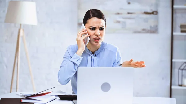 Wütende Geschäftsfrau spricht auf Smartphone, während sie im Büro auf Laptop schaut — Stockfoto