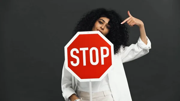 Hispanic woman pointing at signboard with stop lettering isolated on black — Stock Photo